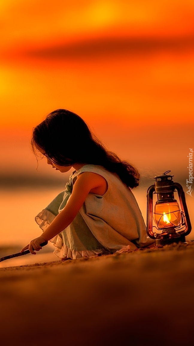 Dziewczynka pisząca patykiem po piasku przy zapalonym lampionie
