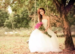 Azjatka w sukni z bukietem kwiatów na huśtawce pod drzewem