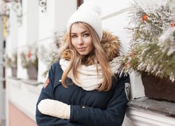 Blondynka w zimowej kurtce i szaliku na tle świątecznej dekoracji w oknie