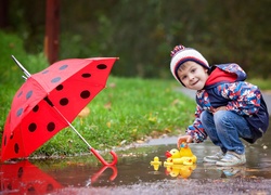 Czerwony parasol i chłopiec bawiący się gumowymi kaczuszkami w kałuży
