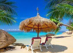Dwa leżaki pod słomianym parasolem i palmami obok głazu na plaży