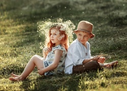 Dziewczynka w wianku i chłopczyk w kapeluszu siedzą na trawie