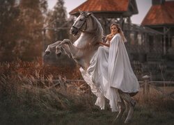 Kobieta w białej pelerynie na siwym koniu