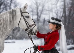 Kobieta w kapeluszu obok siwego konia w zimowej scenerii