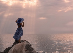 Kobieta w letniej sukience i kapeluszu podziwia morze ze skały