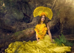 Kobieta w żółtej sukni z wachlarzami we włosach siedzi przy zwalonym drzewie