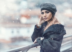 Piękna blondynka w czapce i kożuszku przy barierce w płatkach śniegu