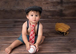 Siedzący chłopczyk w krawacie z piłką i rękawicą do baseballa